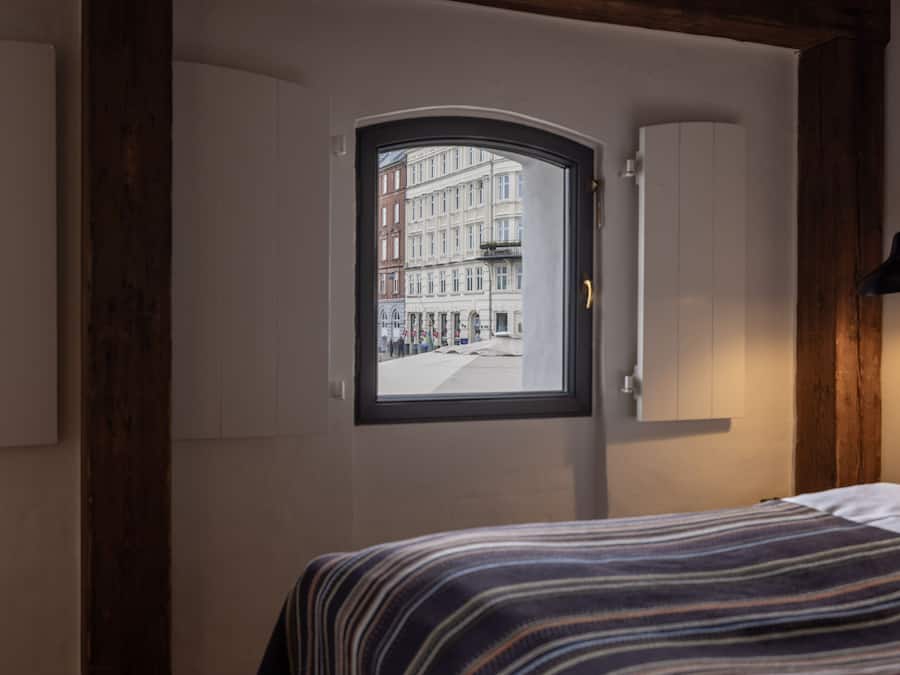 Ett sovrum med ett fönster som vetter ut mot en stadsgata, en säng med ett randigt överkast och ett träpanelat tak.