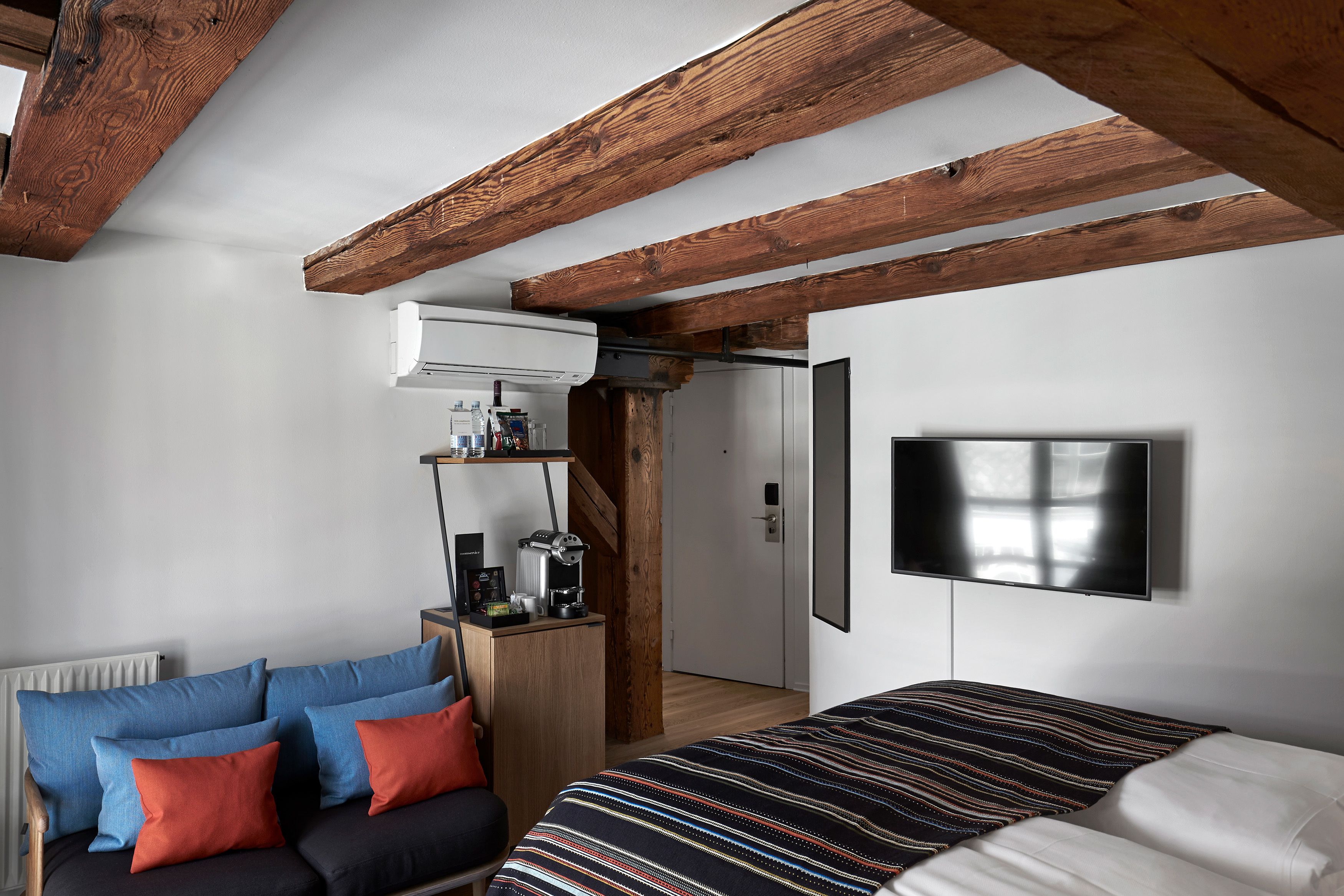 A bedroom with a bed, a sofa, a TV mounted on the wall, and a wooden ceiling.