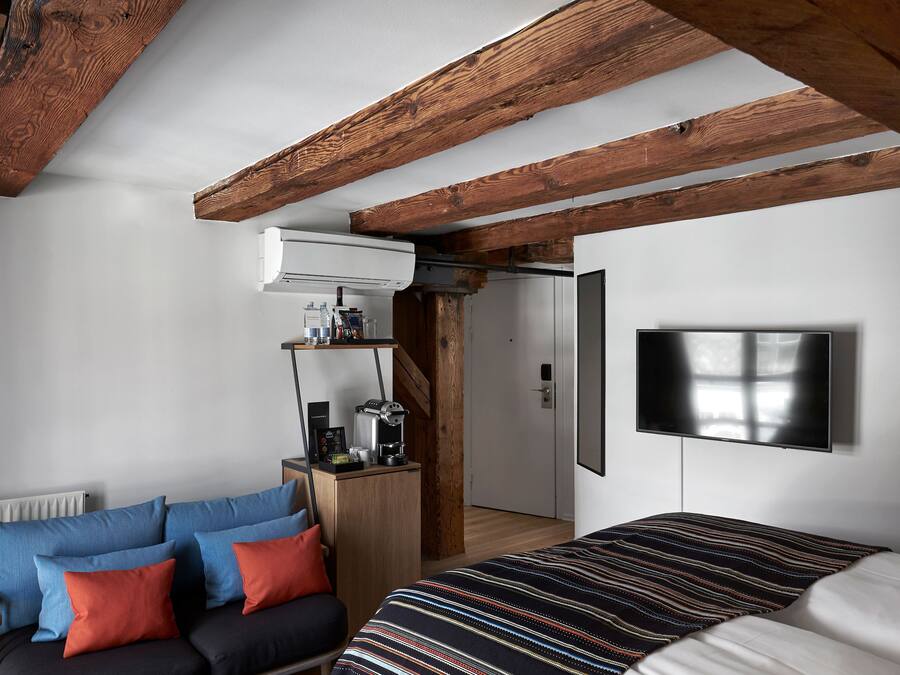A bedroom with a bed, a sofa, a TV mounted on the wall, and a wooden ceiling.