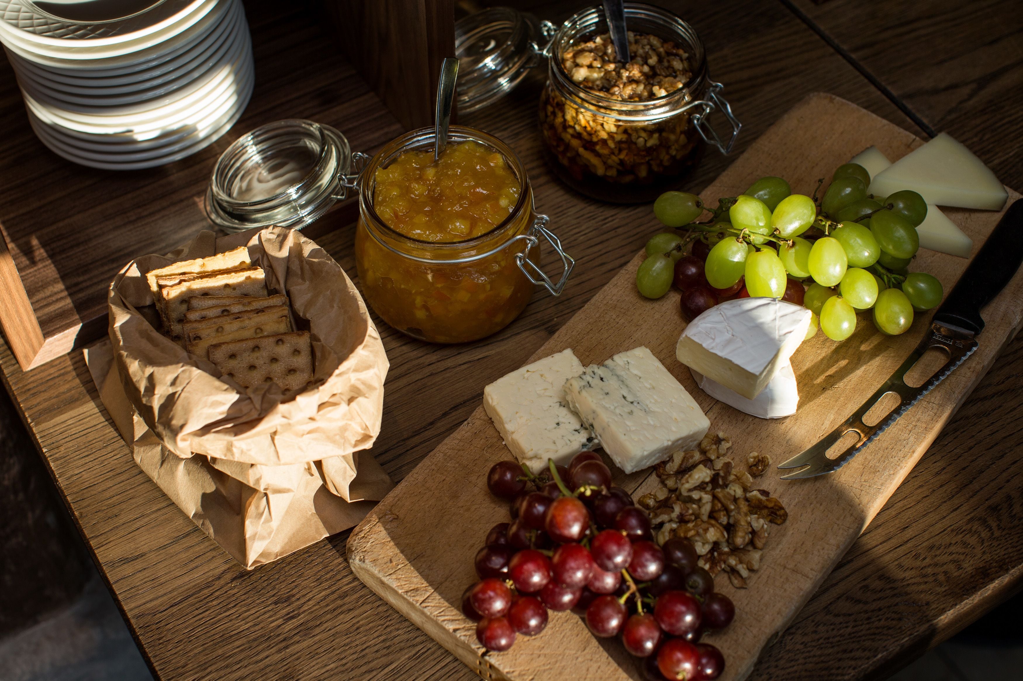Una mesa de madera con diversos alimentos, incluyendo queso, uvas, frutos secos y un tarro de mermelada.