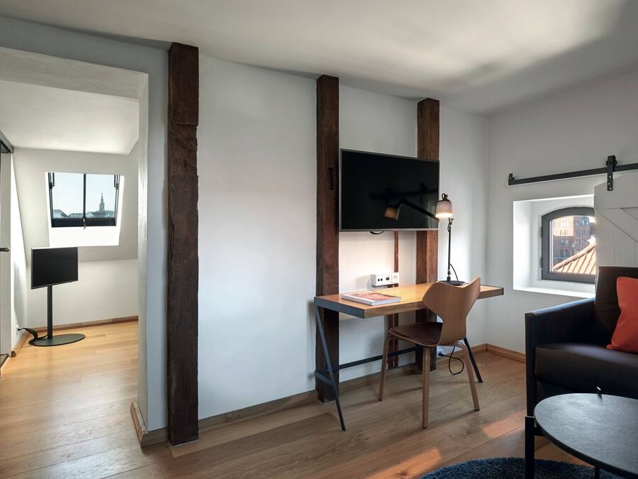 Una habitación moderna con un escritorio de madera, una silla, un televisor de pantalla plana sujeto a una viga de madera y un sofá con un cojín rojo.