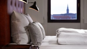 Una cama bien hecha con sábanas blancas, un cabecero y vista a un edificio a través de una ventana.