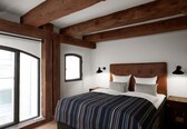 A bedroom with a large window, wooden beams, a bed with striped bedding, and two bedside lamps.