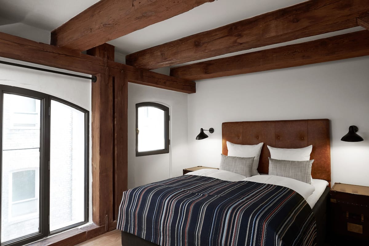 Une chambre avec une grande fenêtre, des poutres apparentes, un lit avec une literie à rayures et deux lampes de chevet.