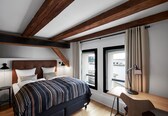 A bedroom with a wooden ceiling, a bed with striped bedding, a desk with a lamp, and a view through a window.