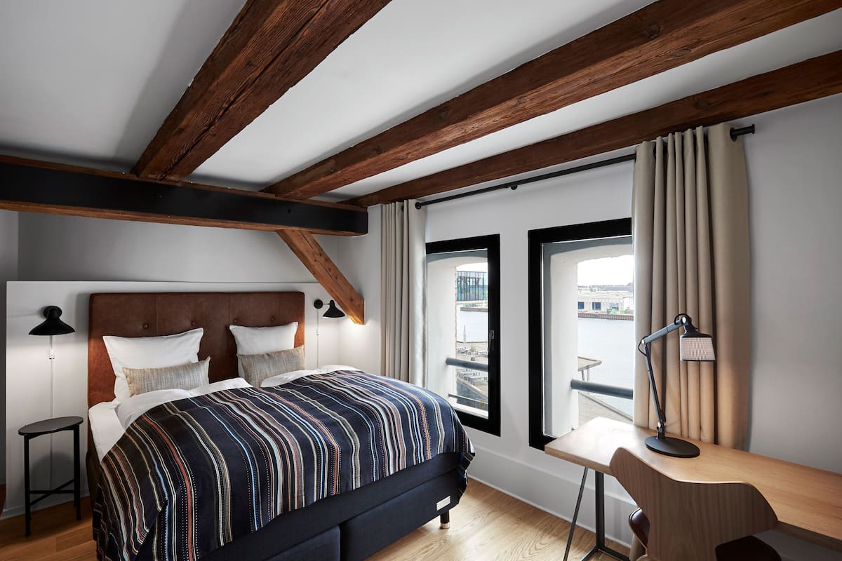 Une chambre avec un plafond en bois, un lit avec une literie à rayures, un bureau avec une lampe et une vue par la fenêtre.