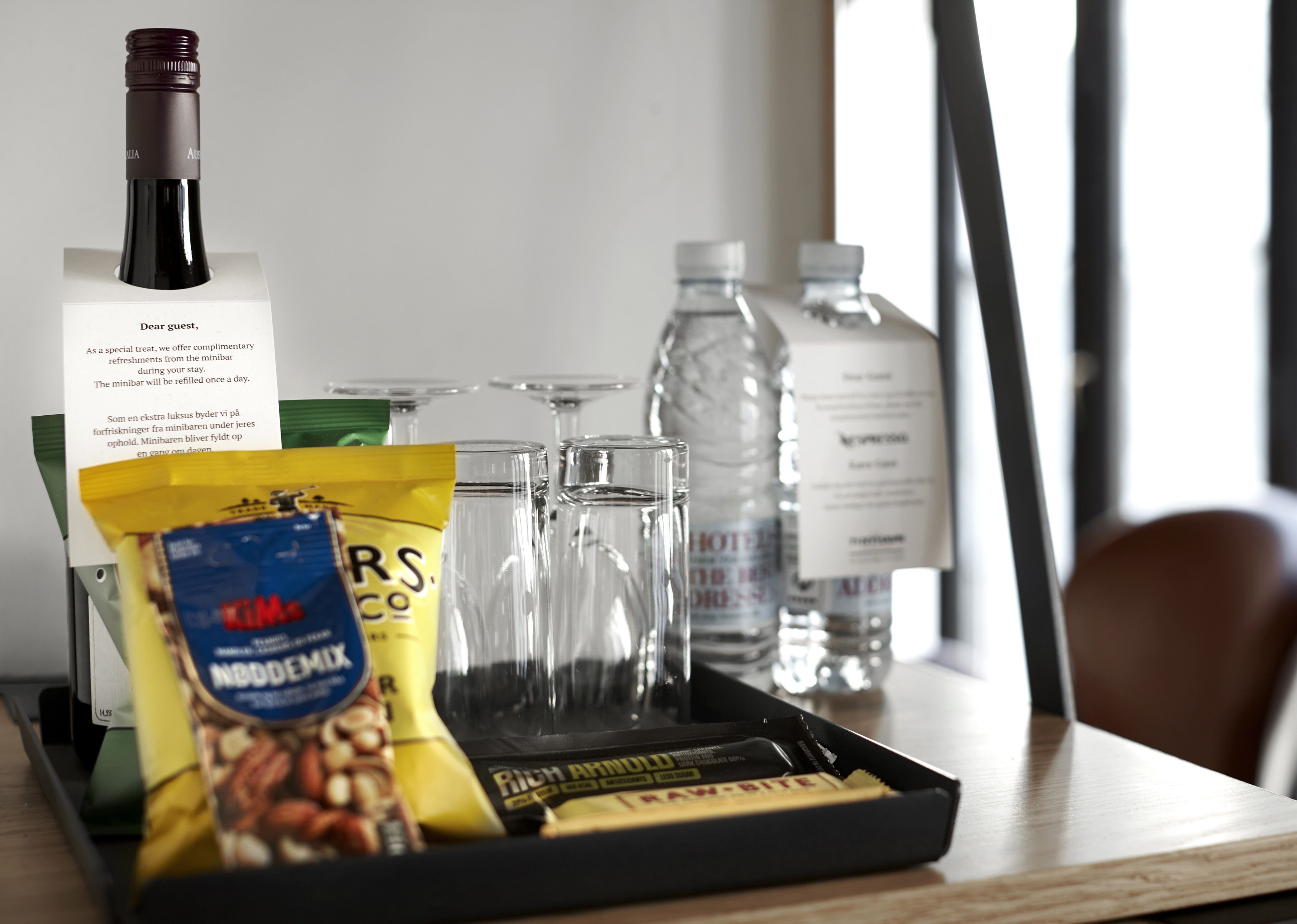 A tray with a bottle of wine, glasses, a water bottle, a bag of nuts, and a chocolate bar.