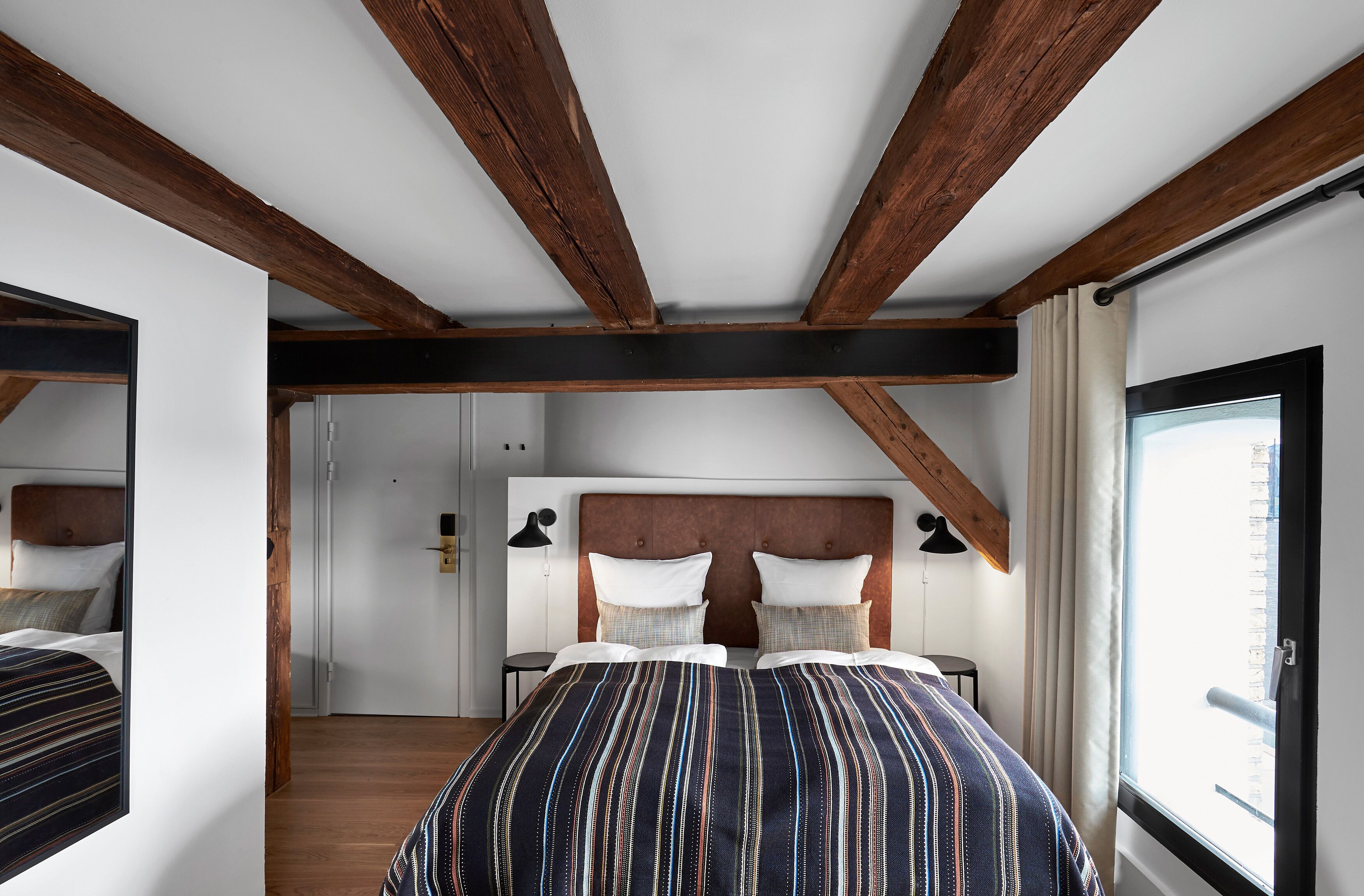 Un dormitorio con una cama grande, vigas de madera en el techo, una ventana con cortinas y una puerta que da a otra habitación.