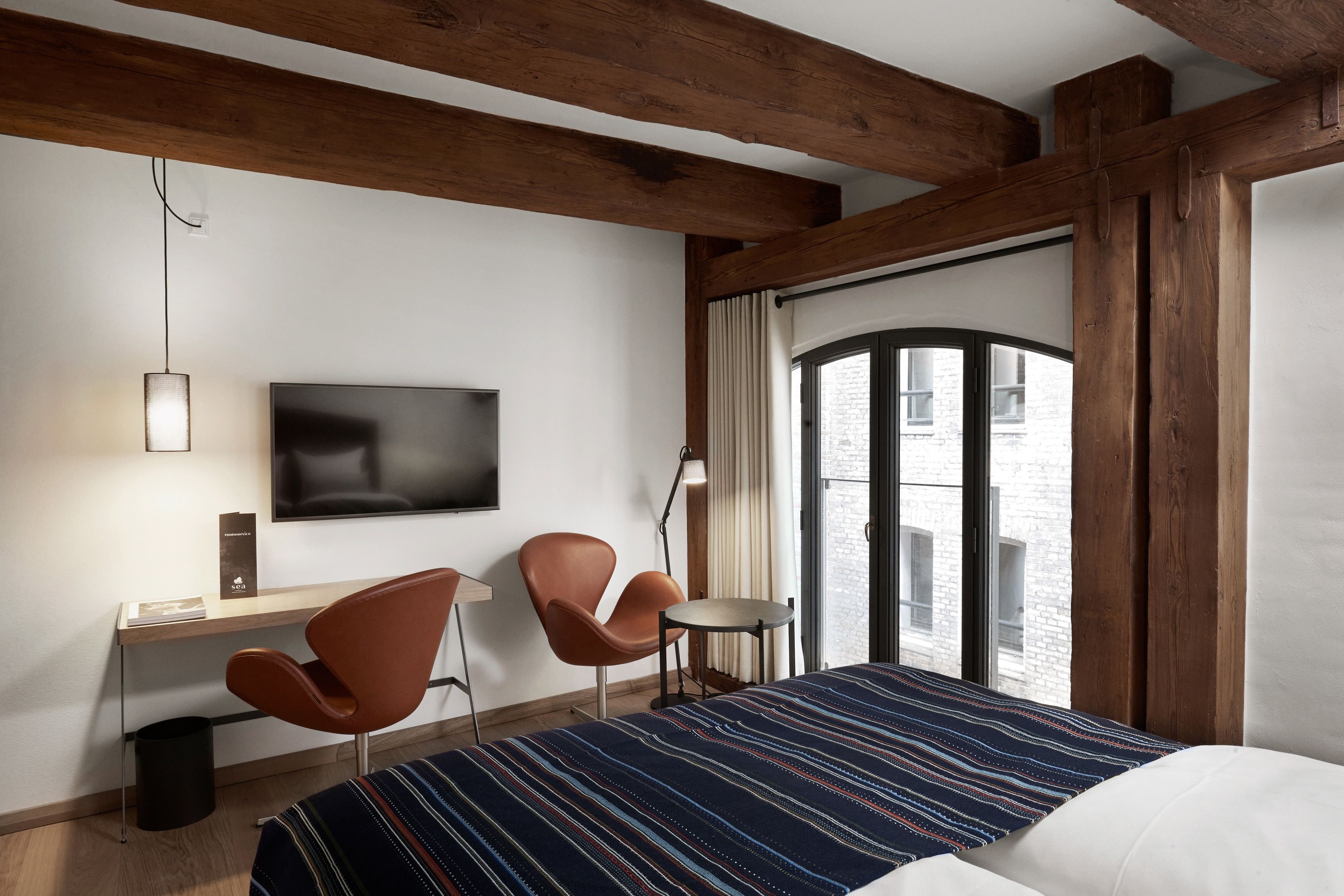 A hotel room with a bed, a desk, two chairs, a TV, and a window with a view of a brick building.