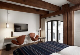A hotel room with a bed, a desk, two chairs, a TV, and a window with a view of a brick building.