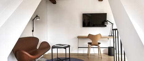 Un dormitorio tipo loft moderno con techo inclinado, un televisor de pantalla plana montado en la pared, un escritorio con silla, un sillón de cuero marrón y una pequeña mesa redonda.
