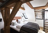 A bedroom with a wooden ceiling structure, a bed with white bedding, a black and white striped blanket, and a view of a brick building through a window.