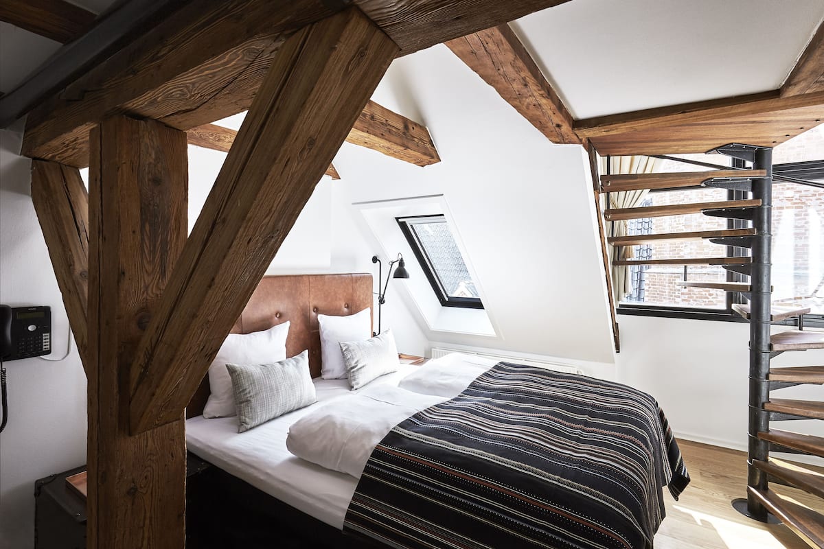 Une chambre avec une structure de plafond en bois, un lit avec une literie blanche, une couverture rayée noire et blanche, et une vue sur un bâtiment en briques par la fenêtre.