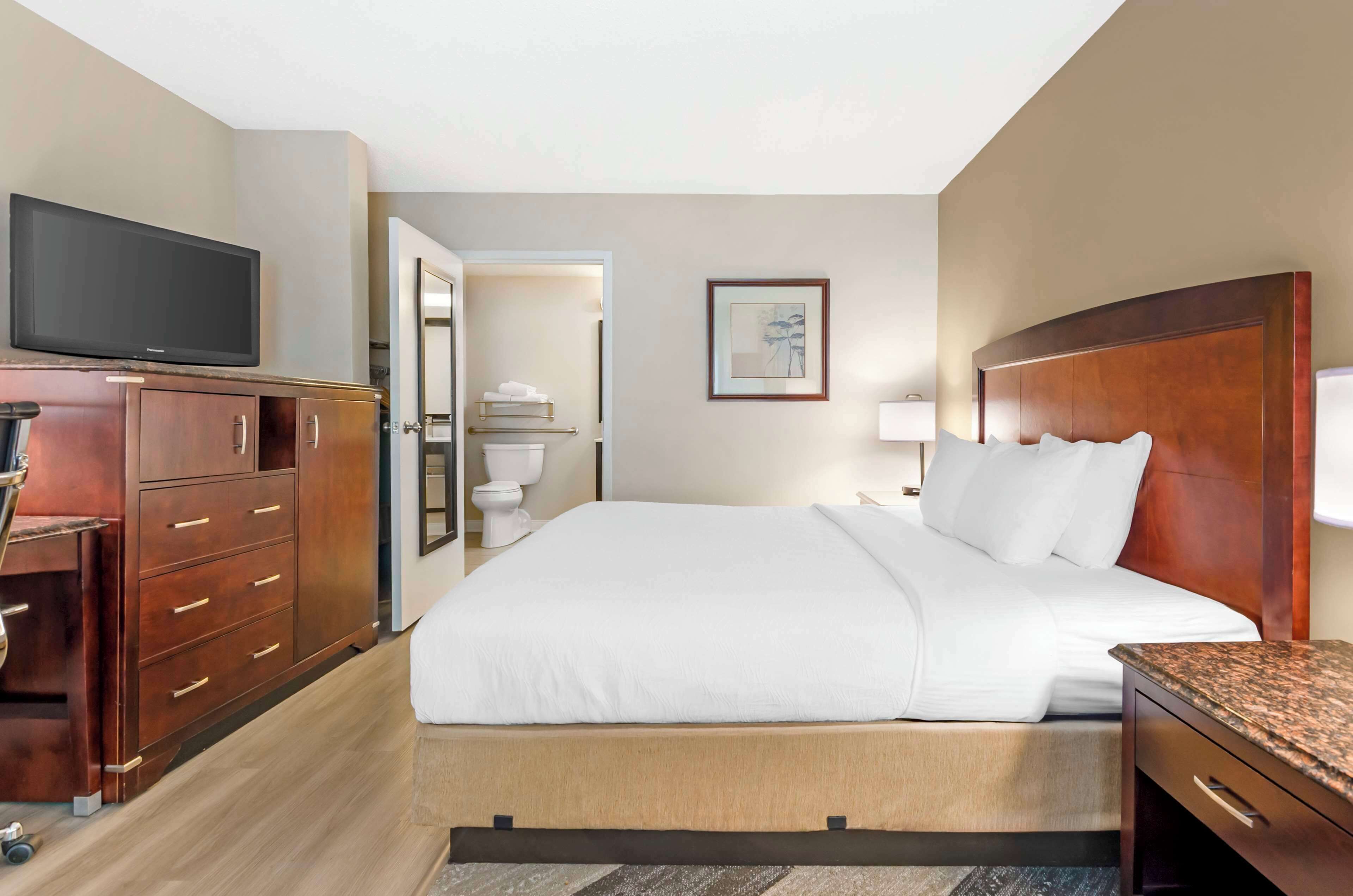 Quarto de hotel com cama, televisão, escrivaninha e banheiro visível através de uma porta aberta.