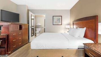 Quarto de hotel com cama, televisão, escrivaninha e banheiro visível através de uma porta aberta.