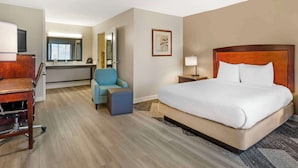 A hotel room with a bed, a desk, a chair, a small table, a lamp, and a painting on the wall.