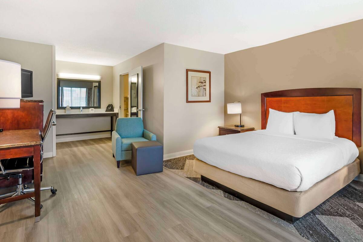 A hotel room with a bed, a desk, a chair, a small table, a lamp, and a painting on the wall.