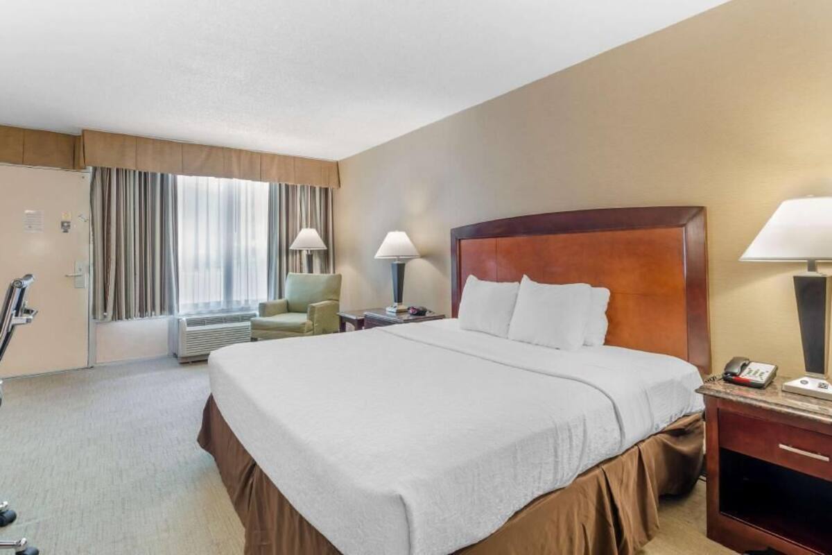 A hotel room with a large bed, a desk with a lamp, a chair, and a window with curtains.