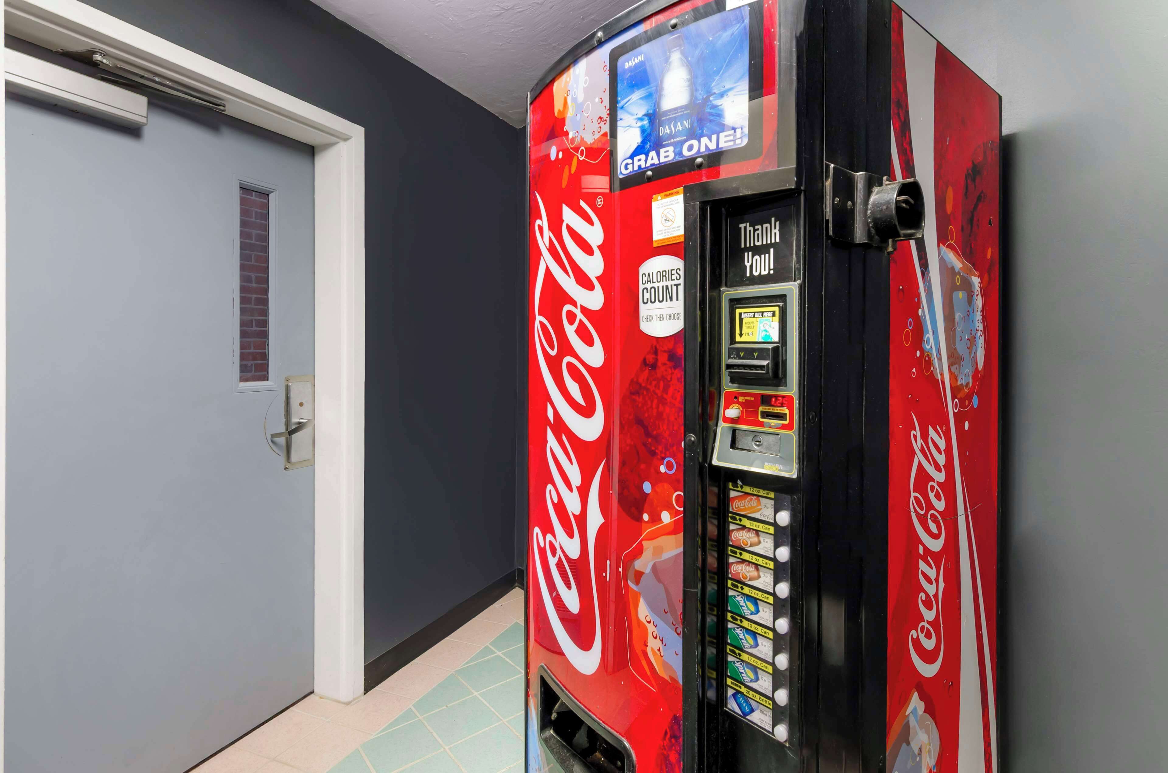 A Coca-Cola vending machine in a hallway with a door and a window.