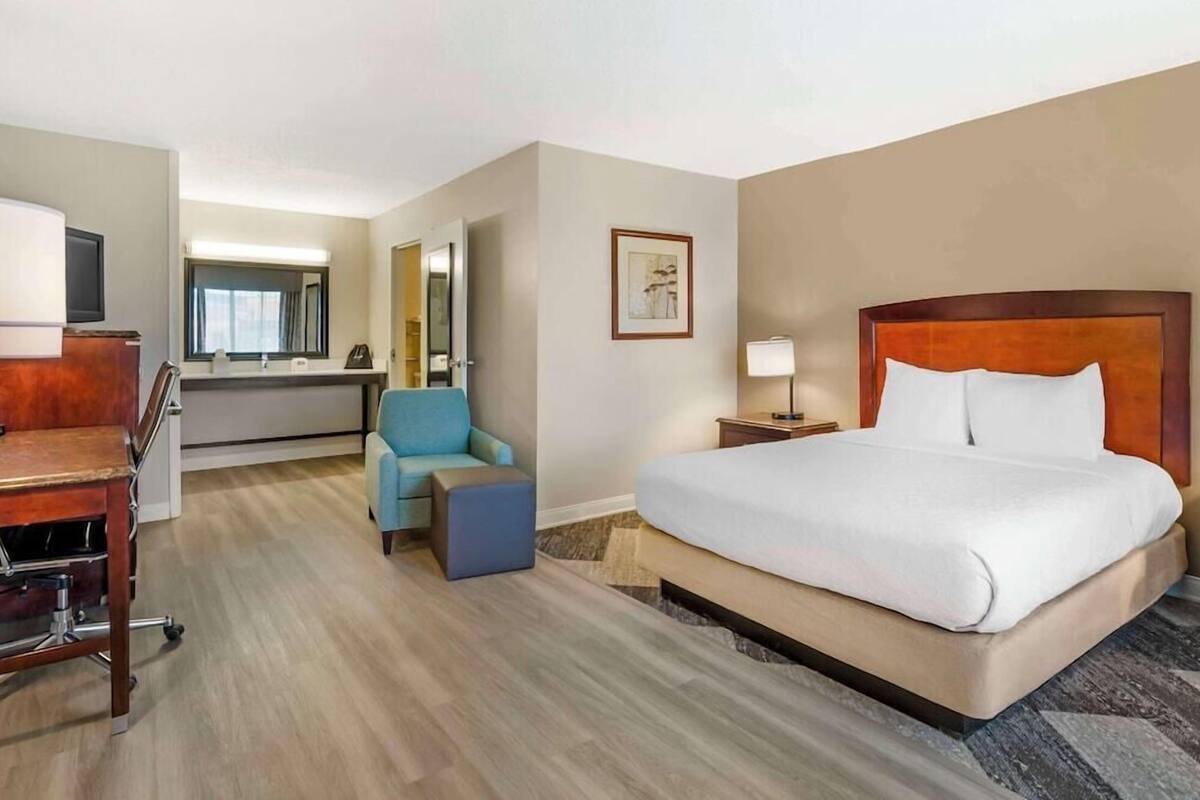 A hotel room with a bed, desk, chair, and a small ottoman.