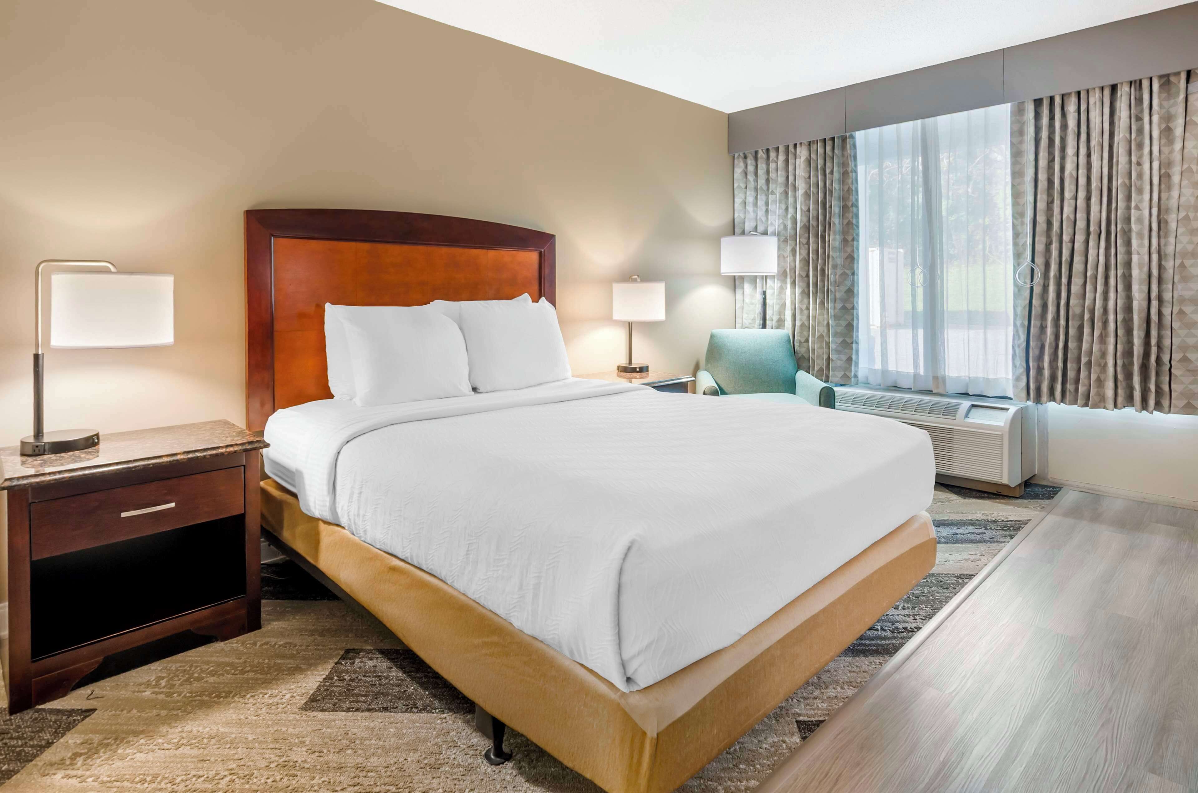 A hotel room with a large bed, two bedside tables with lamps, a chair, a large window with curtains, and a wall-mounted air conditioner.