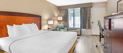 A hotel room with a large bed, a desk with a lamp, a chair, a dresser, and a window with curtains.