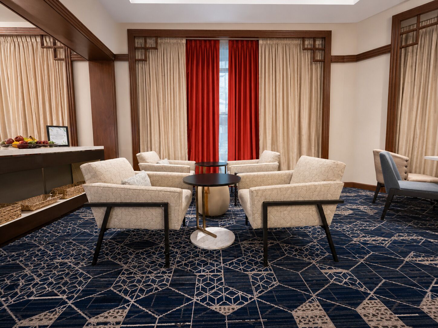 Ett hotellrum med ett geometriskt mönstrat mattgolv, ett runt bord, fyra fåtöljer och ett stort fönster med röda gardiner.