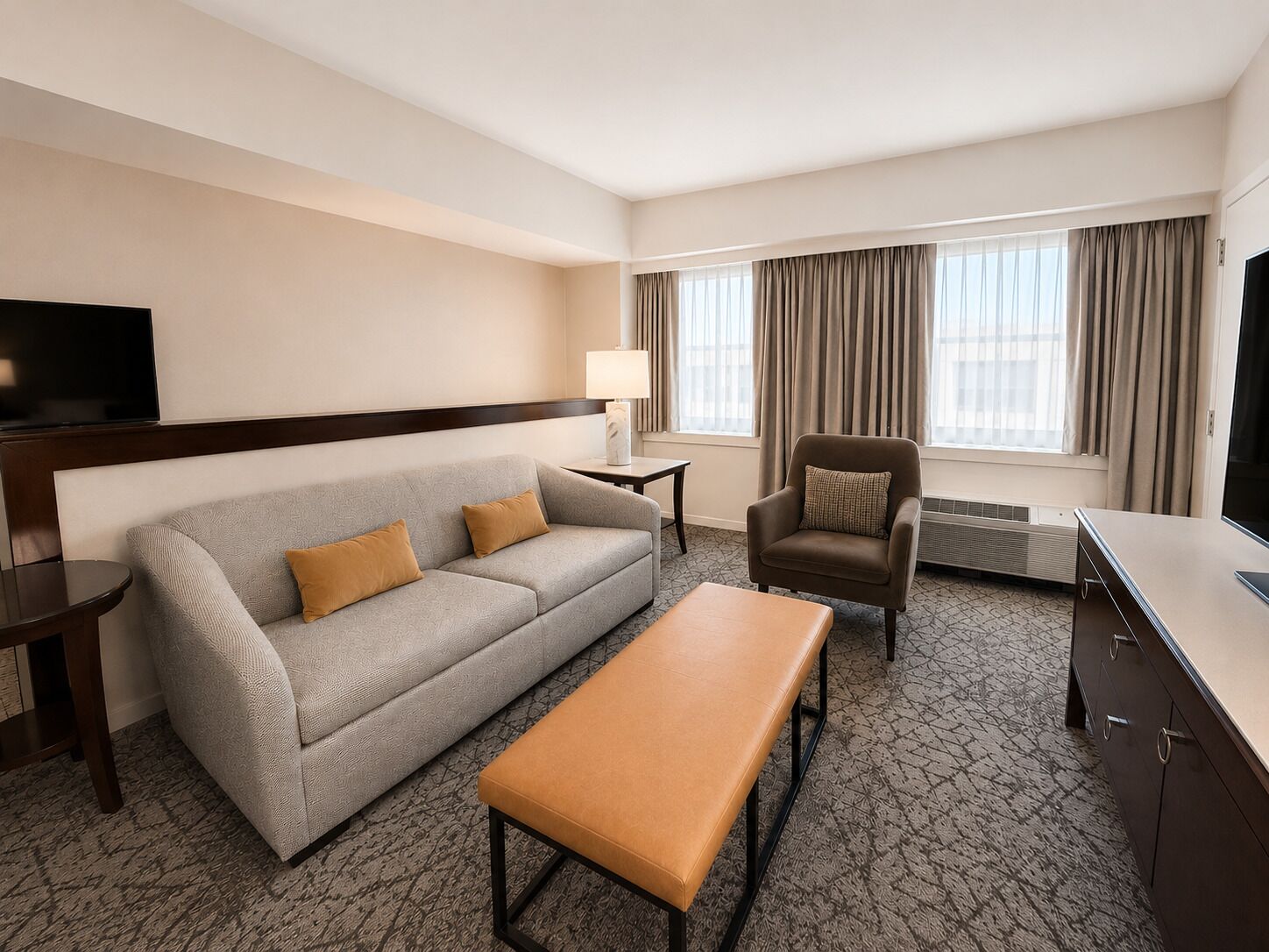A hotel room with a sofa, armchair, bench, TV, lamp, and air conditioning unit.