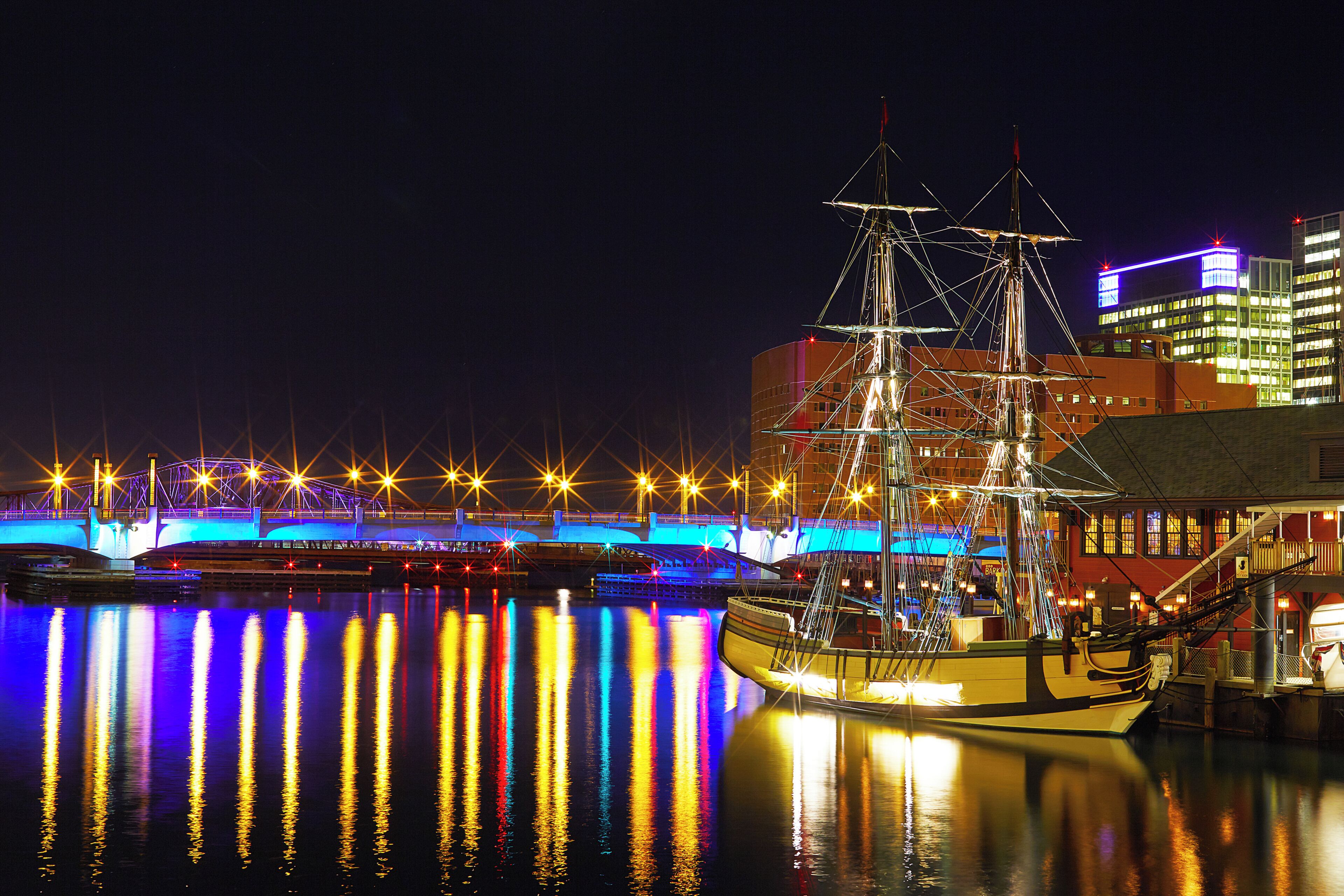 A historic ship docked at a waterfront with illuminated buildings in the background.