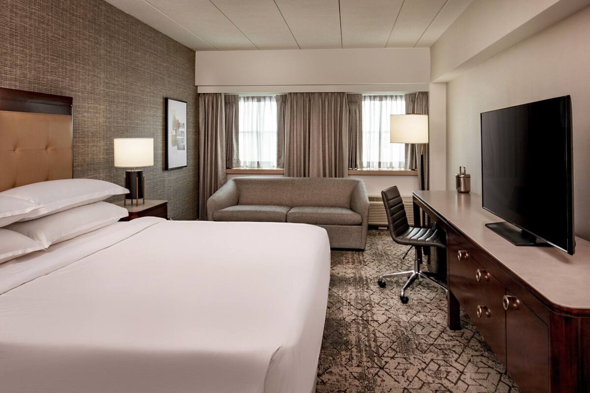 A hotel room with a large bed, a sofa, a desk with a TV, and a window with curtains.