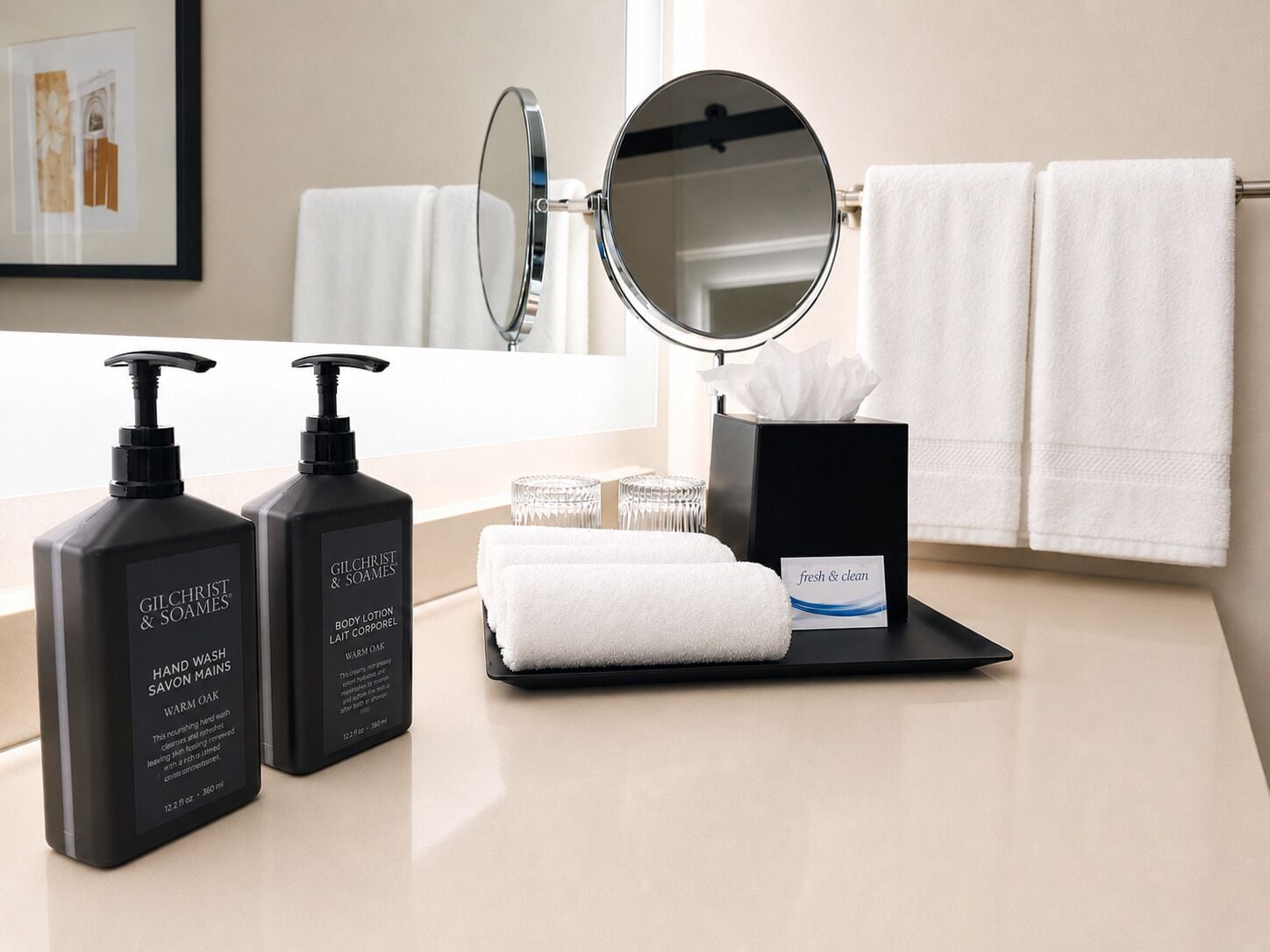 A bathroom with a mirror, two bottles of Gilchrist & Soames body lotion, a tissue box, and a folded towel.