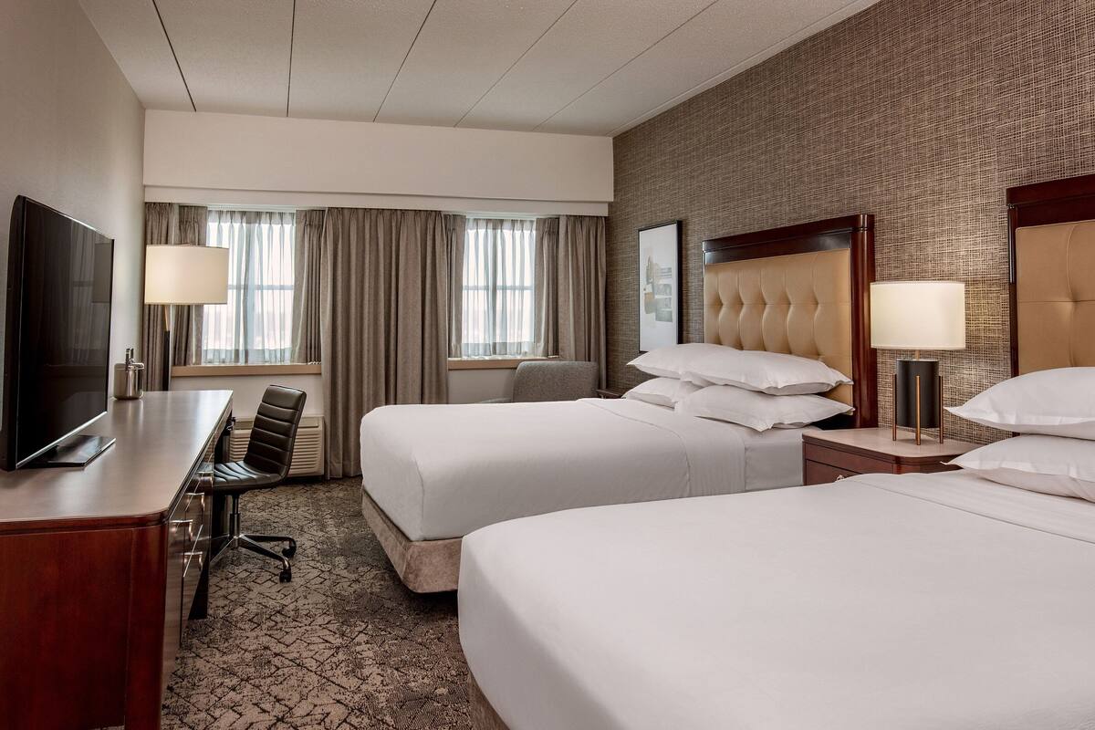 A hotel room with two beds, a desk, a TV, and a large window with curtains.