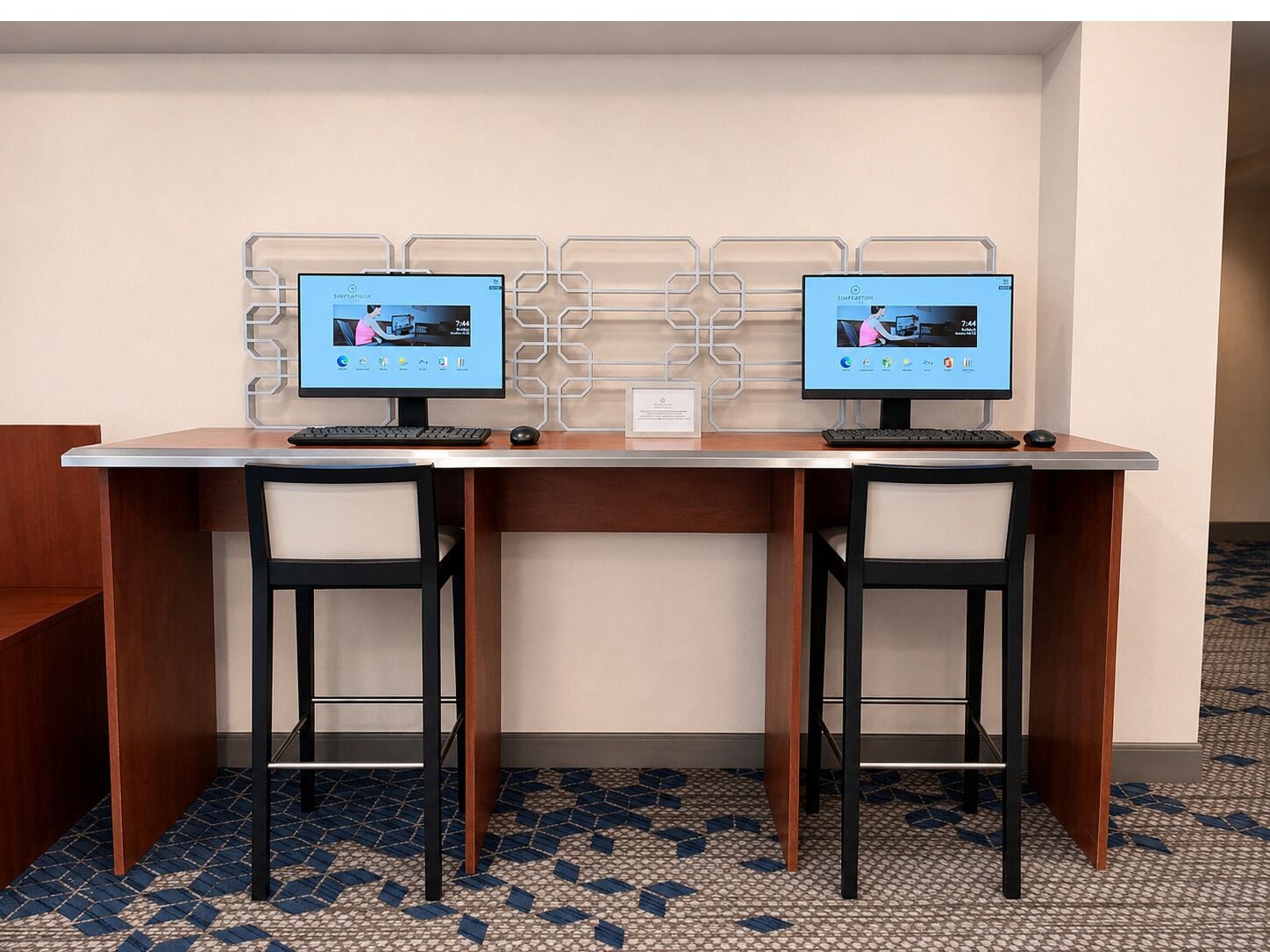 A hotel workspace with two computer monitors, a wooden desk, two black chairs, and a patterned floor.