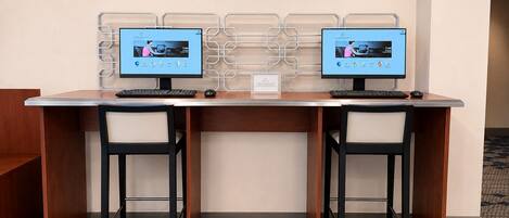 A hotel workspace with two computer monitors, a wooden desk, two black chairs, and a patterned floor.