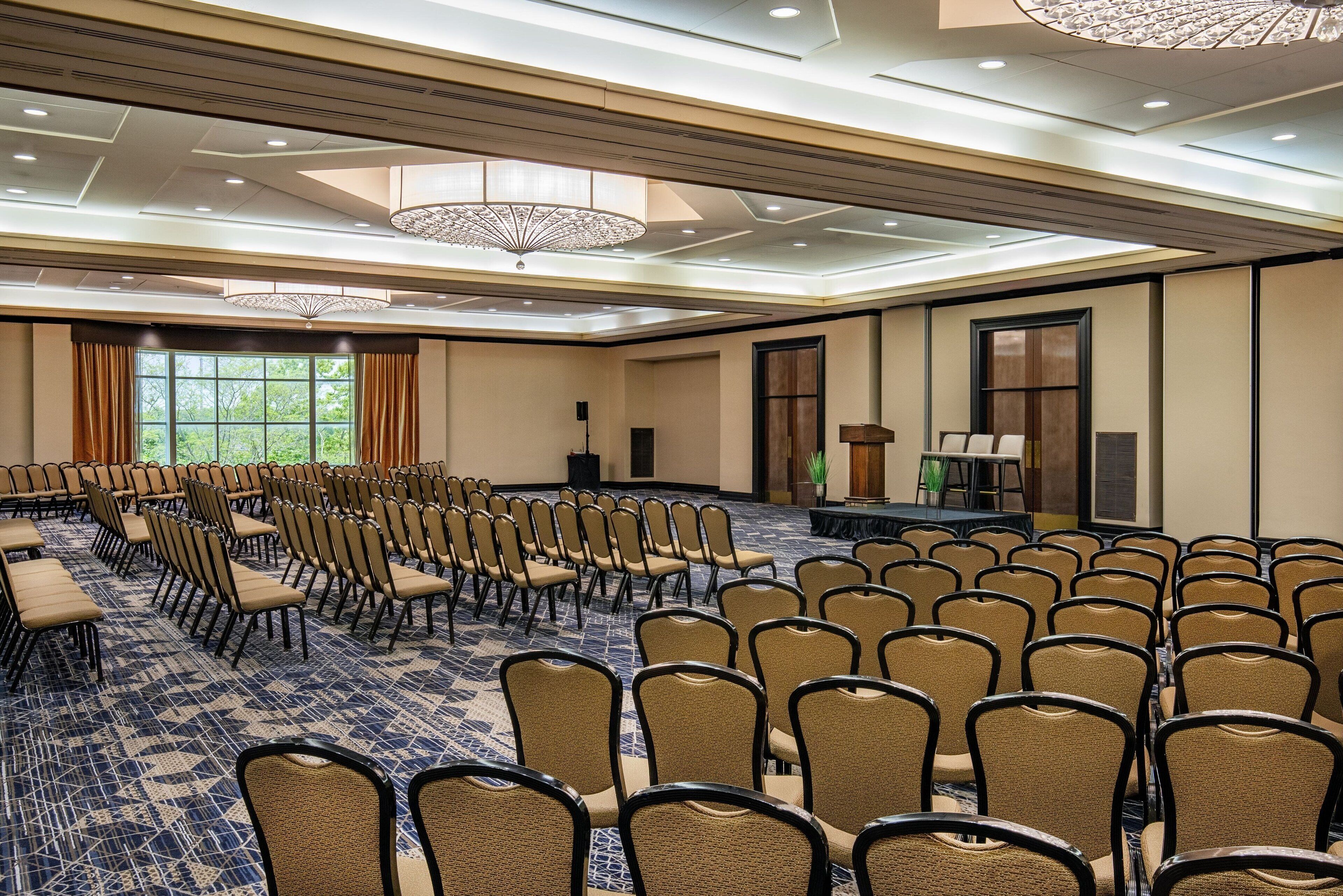 A conference room with rows of chairs, a stage, and a large window with curtains.