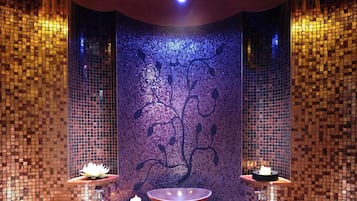 Relax in a serene spa room with a central sink and two seating areas, surrounded by mosaic tile walls and a stylish ceiling light fixture.