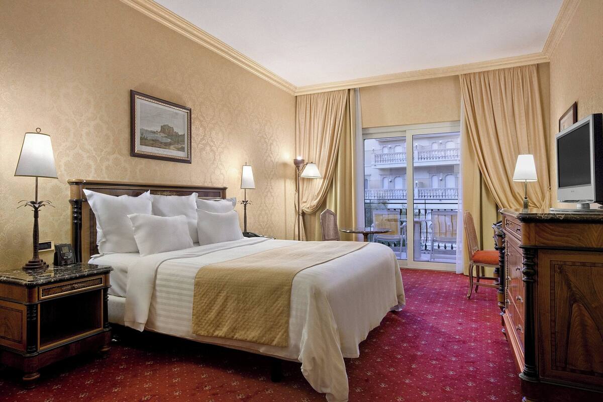 A hotel room with a large bed, two bedside tables, a dresser, a television, and a balcony with a railing.