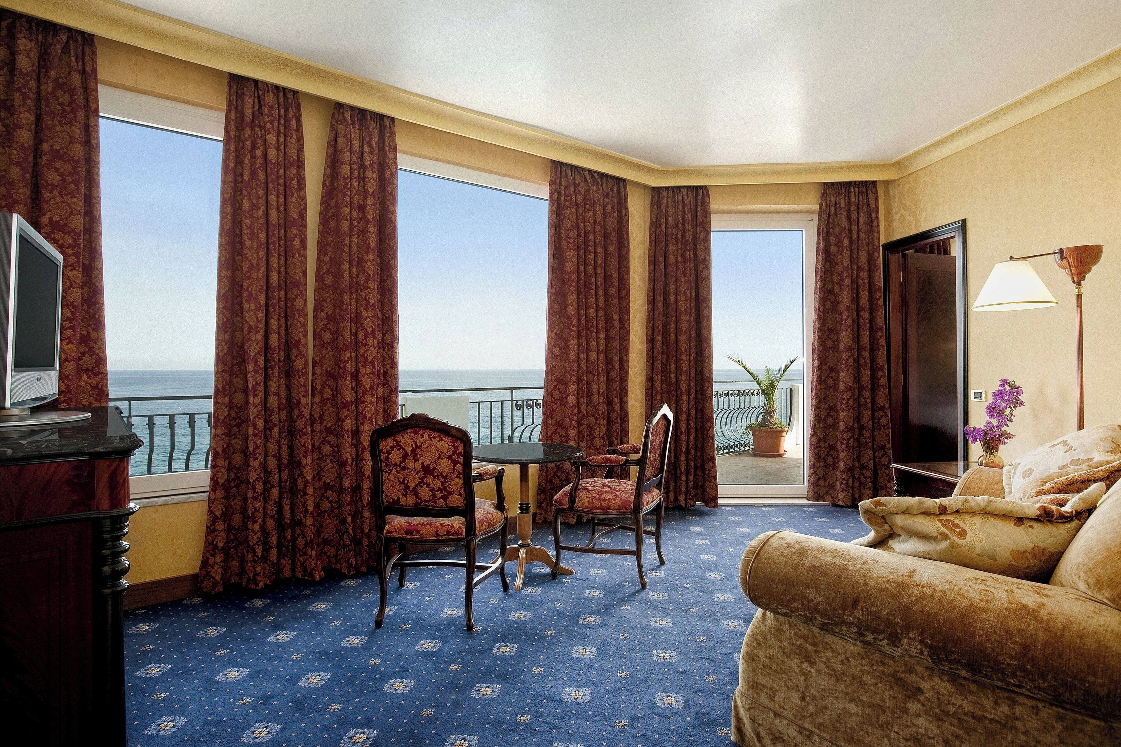 A hotel room with a large window, a TV, a sofa, a table with chairs, and a lamp.