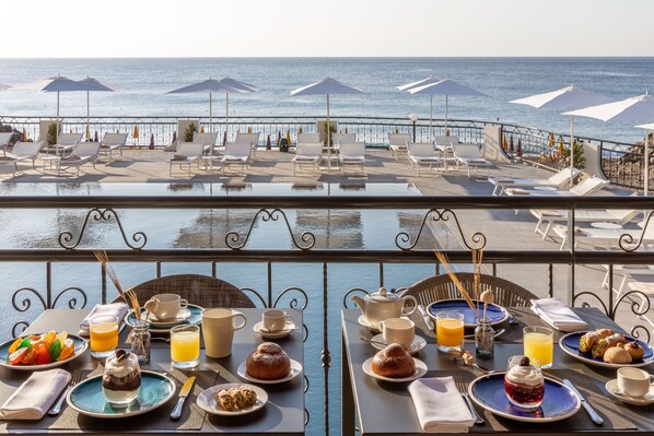 Terraza con mesas dispuestas para el desayuno con vistas al mar, que incluye sillones de sol y sombrillas.