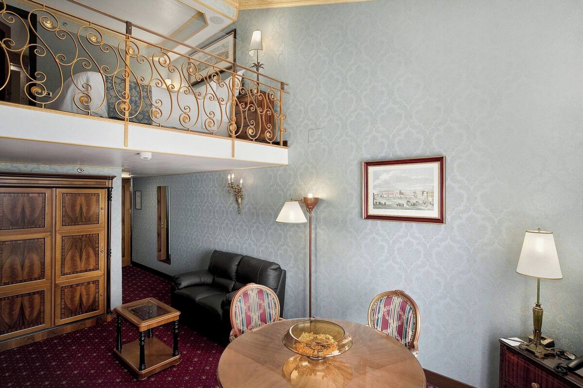 A room with a high ceiling, a balcony with ornate railings, a dining table with a glass centerpiece, a black leather sofa, patterned chairs, and a framed picture on the wall.