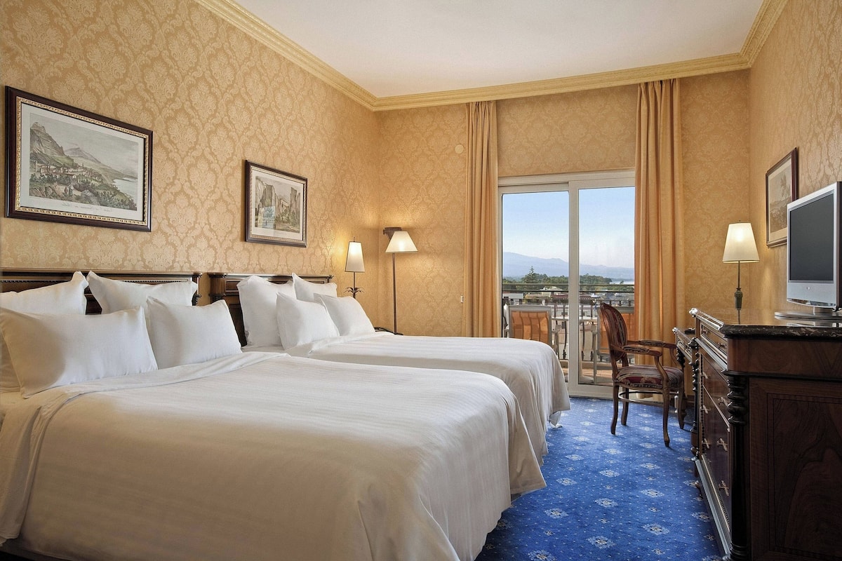 A hotel room with two beds, a television, a desk, and a view of the outdoors.