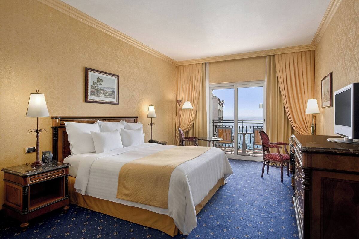 A hotel room with a large bed, two bedside tables with lamps, a TV, a desk with a chair, and a balcony with a view.
