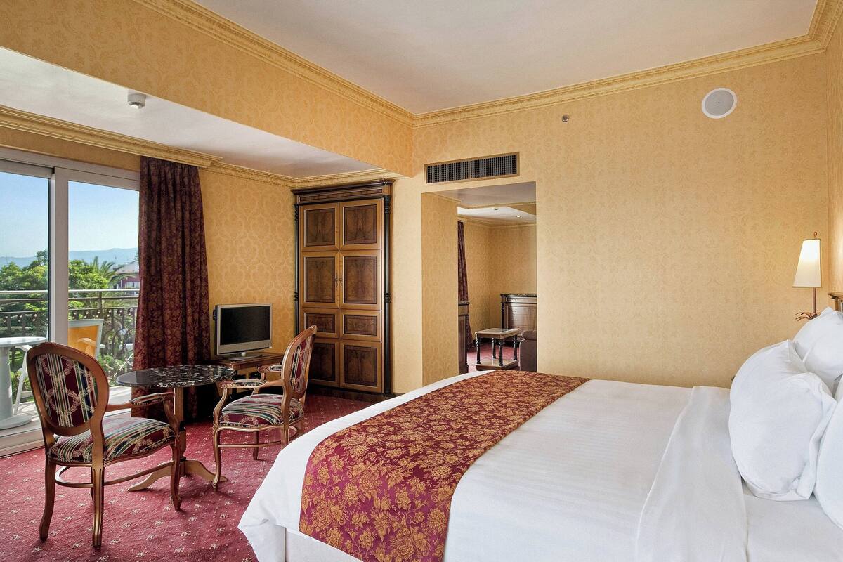 A hotel room with a large bed, a desk with a chair, a television, and a balcony with curtains.