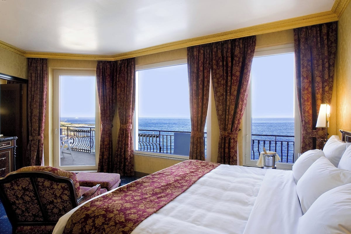 A hotel room with a large bed, a chair, a desk, and a view of the ocean through large windows.