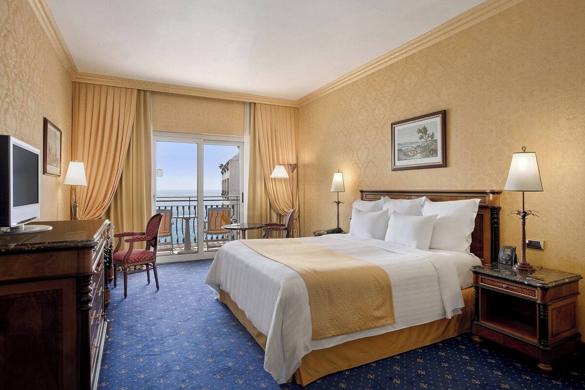 A hotel room with a large bed, a desk with a TV, a chair, a small table, and a balcony with curtains.