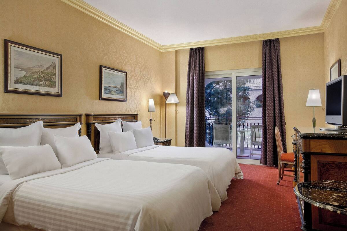 A hotel room with two beds, a balcony with a view, and a desk with a television.
