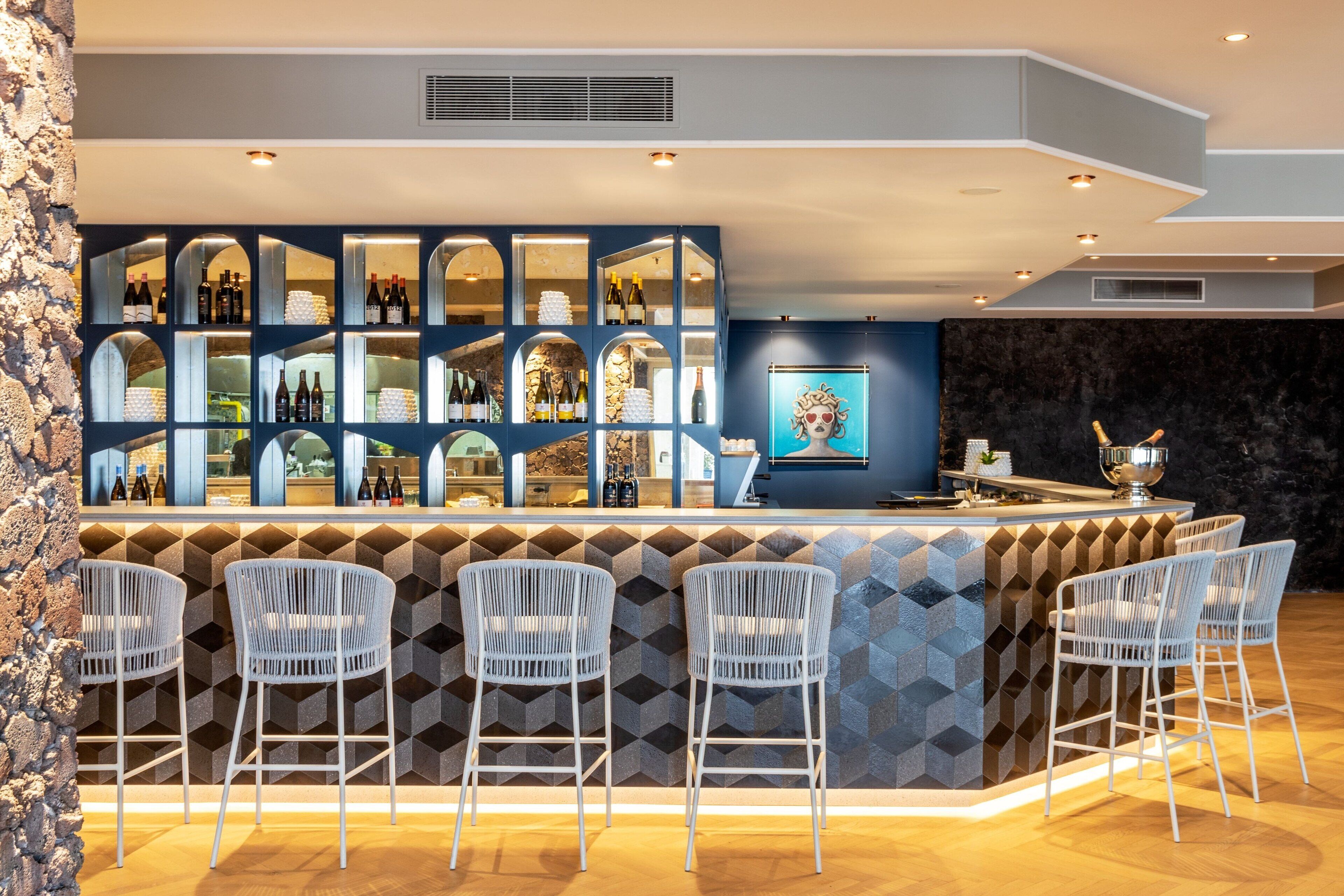 Un espace bar au design moderne, avec un comptoir doté de plusieurs étagères où sont exposées des bouteilles, et trois tabourets de bar blancs.