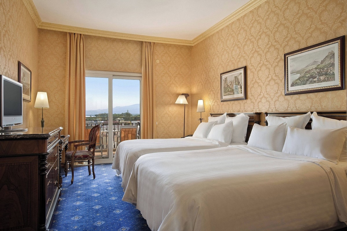 A hotel room with two beds, a desk with a television, and a view of a mountainous landscape through a large window.