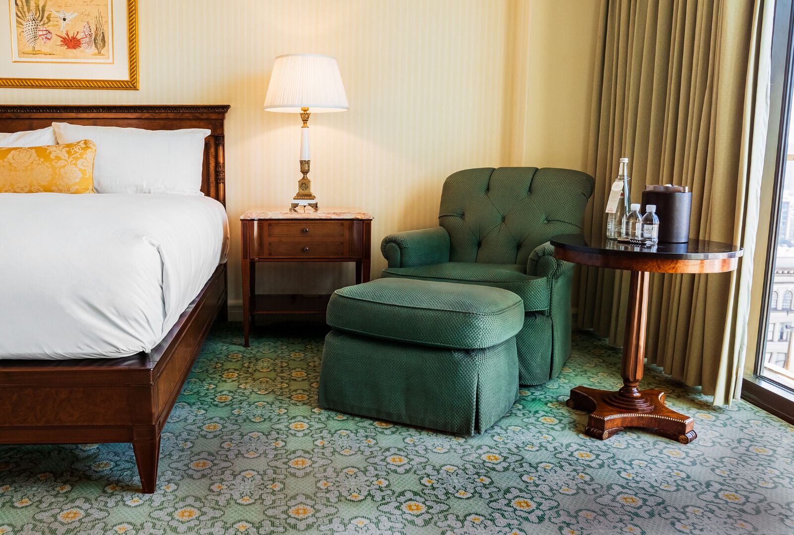 Une chambre d’hôtel avec un lit, un fauteuil vert, une petite table avec une lampe et un tapis à motifs.