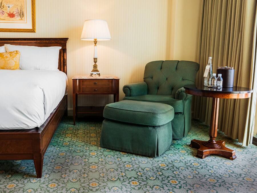 Une chambre d’hôtel avec un lit, un fauteuil vert, une petite table avec une lampe et un tapis à motifs.